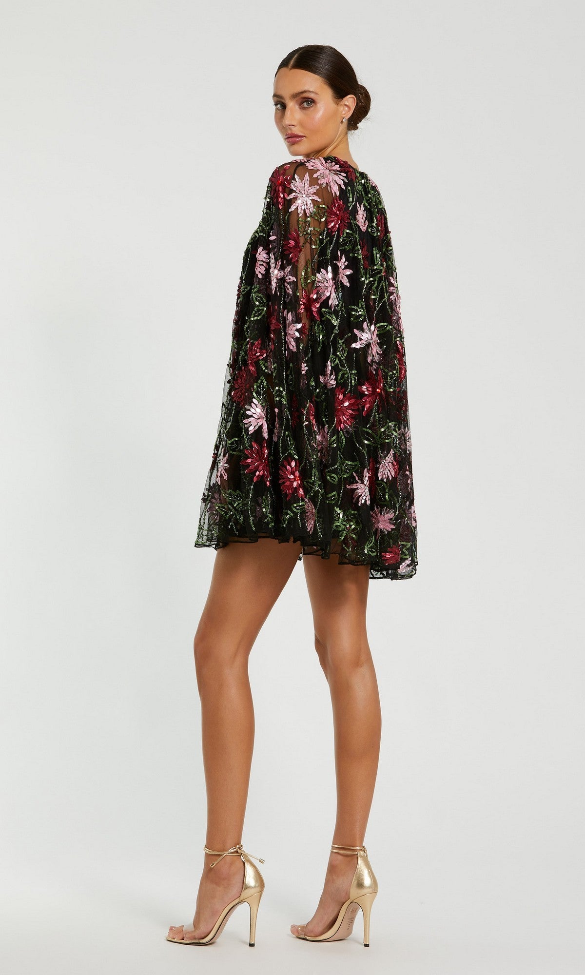 A woman in profile wears the Mac Duggal 6123 Sequin Short Wedding Guest Dress—black with colorful floral embroidery—and gold high-heeled sandals, posed against a plain white background.
